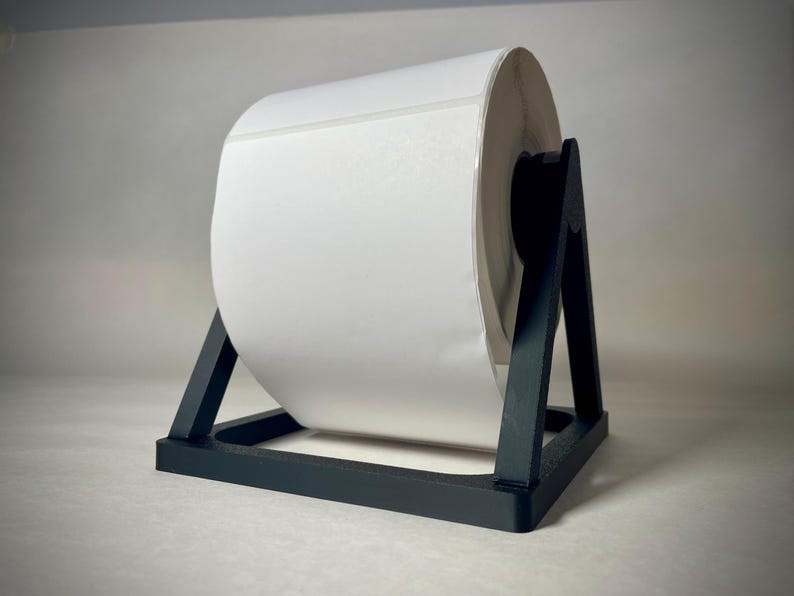 May include: A large roll of white labels sits on a black label holder. The label roll is centered in the frame, with the black holder providing support. The background is a neutral gray.