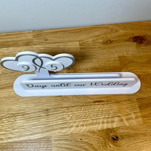 May include: White wooden countdown sign with two gray hearts and the text "Days until our Wedding".