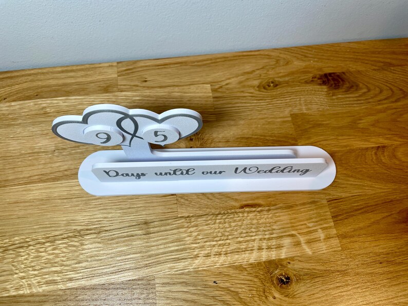 May include: White wooden countdown sign with two gray hearts and the text "Days until our Wedding".