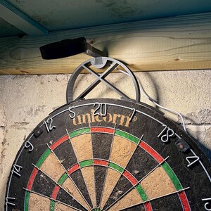 Dart board with attached lamp on the top. The lamp extends out in front of the board in order to cast light on the play area. Lamp is black modern design with a puck light at the end. With the lamp on.