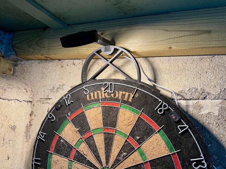 Dart board with attached lamp on the top. The lamp extends out in front of the board in order to cast light on the play area. Lamp is black modern design with a puck light at the end. With the lamp on.