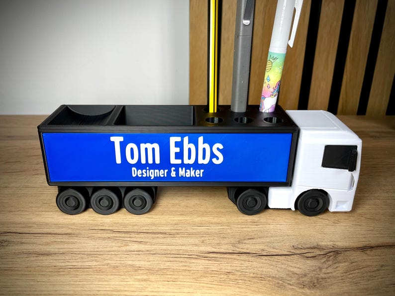 Pu&ograve; includere: Organizzatore da scrivania nero e bianco a forma di camion. Il pannello laterale blu mostra il testo "Tom Ebbs Designer & Maker" in bianco. Contiene penne, matite e altri articoli per l'ufficio.