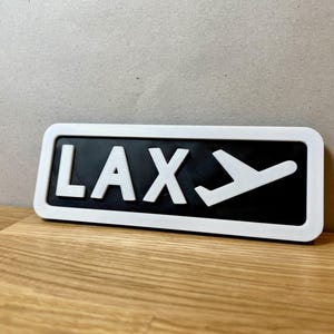 May include: White sign with black background and white lettering that reads "LAX" with a white airplane icon next to it.