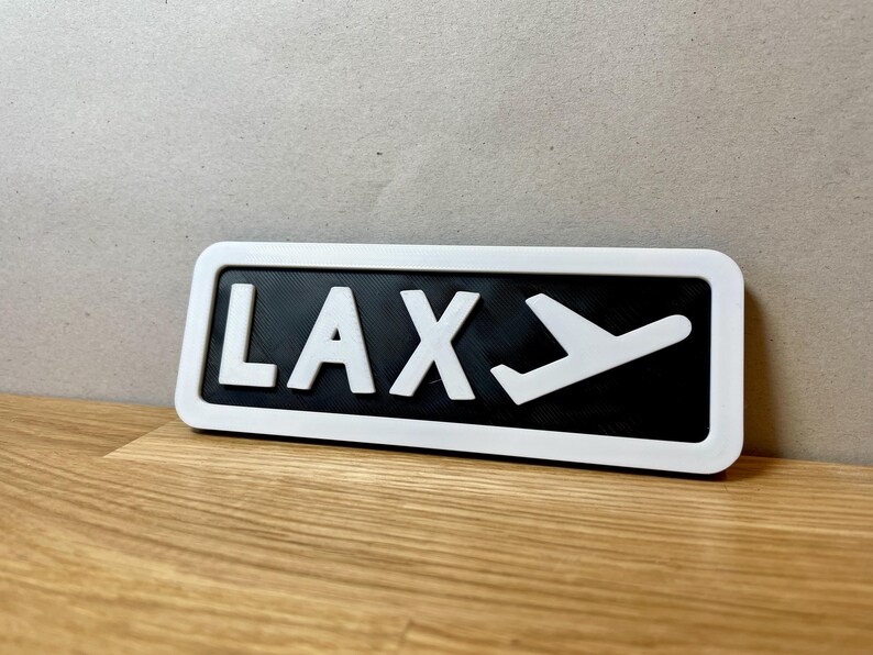 May include: White sign with black background and white lettering that reads "LAX" with a white airplane icon next to it.