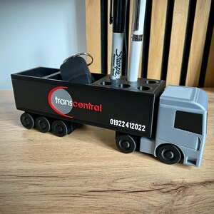 Pu&ograve; includere: Un organizer da scrivania nero e grigio a forma di camion. Dispone di scomparti per penne e altri piccoli oggetti. Il logo "transcentral" e il numero di telefono 01922 412022 sono visualizzati sul lato.