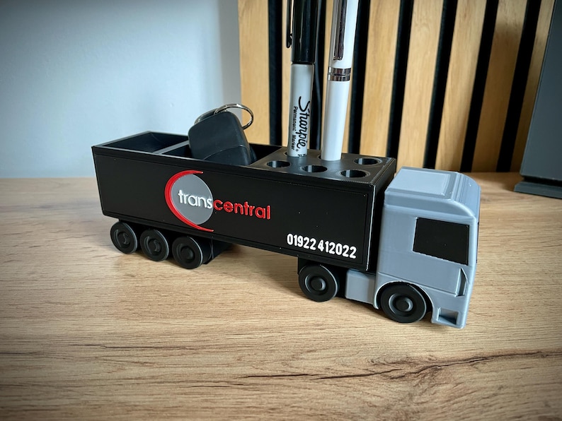 Pu&ograve; includere: Un organizer da scrivania nero e grigio a forma di camion. Dispone di scomparti per penne e altri piccoli oggetti. Il logo "transcentral" e il numero di telefono 01922 412022 sono visualizzati sul lato.