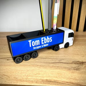 Pu&ograve; includere: Un organizer da scrivania bianco e nero a forma di camion. Il pannello laterale blu mostra il testo "Tom Ebbs Designer & Maker" in bianco. Contiene penne e matite, tra cui una matita gialla e una penna multicolore.