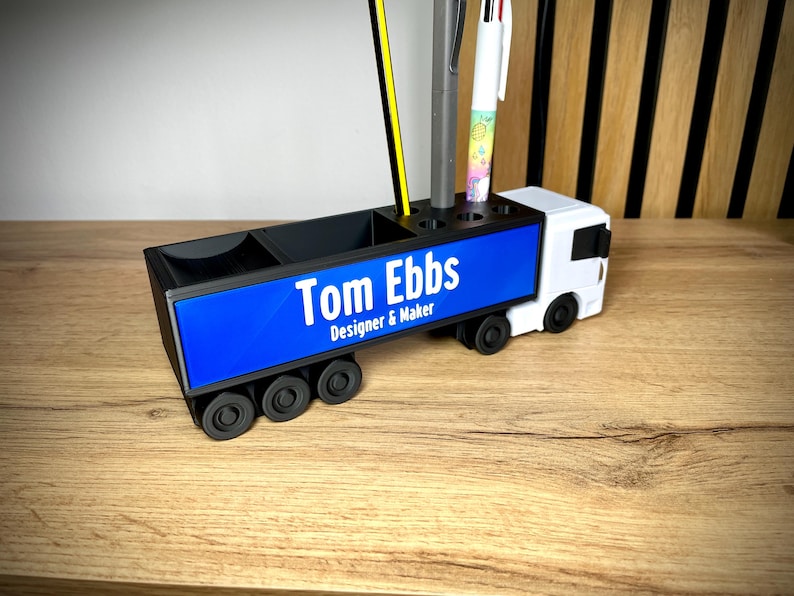 Pu&ograve; includere: Un organizer da scrivania bianco e nero a forma di camion. Il pannello laterale blu mostra il testo "Tom Ebbs Designer & Maker" in bianco. Contiene penne e matite, tra cui una matita gialla e una penna multicolore.