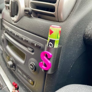 Pu&ograve; includere: Un porta cellulare per auto in plastica nera con una lettera "S" rosa stampata. Un tubo di balsamo per labbra verde e bianco &egrave; inserito nel supporto.