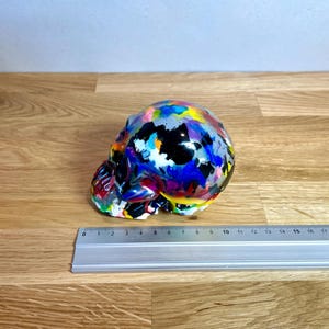 May include: A colourful skull sculpture with a black base and splashes of paint in various colours, including blue, pink, yellow, and white.
