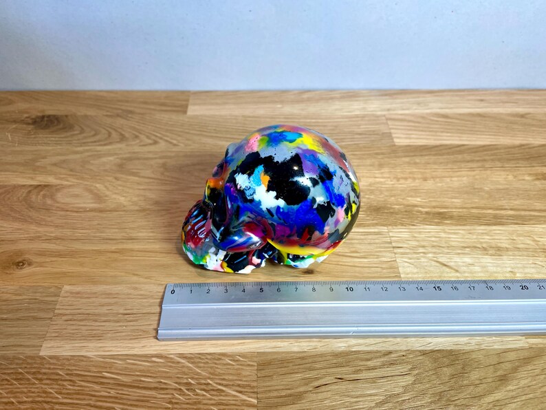 May include: A colourful skull sculpture with a black base and splashes of paint in various colours, including blue, pink, yellow, and white.