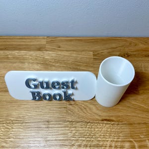 pen pot on the right with a sign that says guest book. Shown on a wooden desk with grey background. White pen pot with a white sign with silver text.