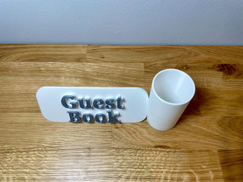 pen pot on the right with a sign that says guest book. Shown on a wooden desk with grey background. White pen pot with a white sign with silver text.