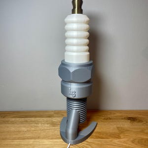 Oversized spark plug lamp with the white ceramic section designed as a light shade. Bronze top section, white shade and silver base. With a white cable. Stood on a wooden desk and grey background.