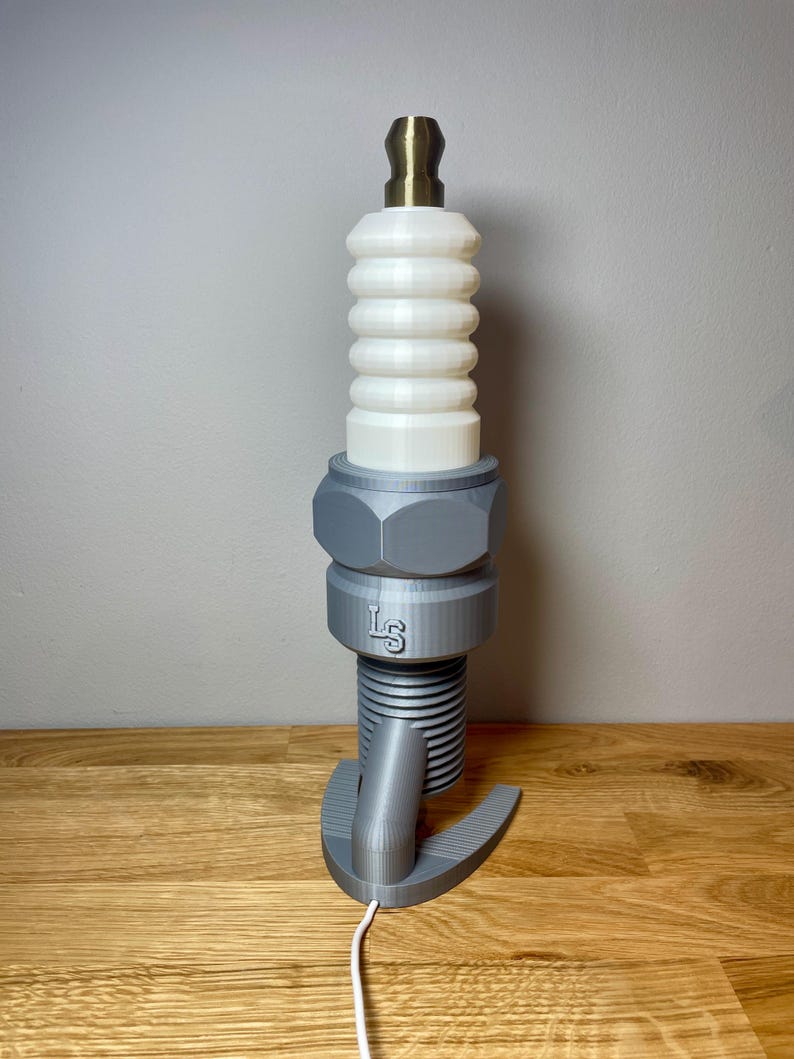 Oversized spark plug lamp with the white ceramic section designed as a light shade. Bronze top section, white shade and silver base. With a white cable. Stood on a wooden desk and grey background.