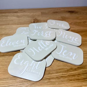 May include: A set of 11 beige square cards with white text. The cards feature different phrases and numbers, such as "One Month", "First", "Hello World", "Eight Months", "Eleven Months", "Nine Months", "Ten Months", and "Two Months".