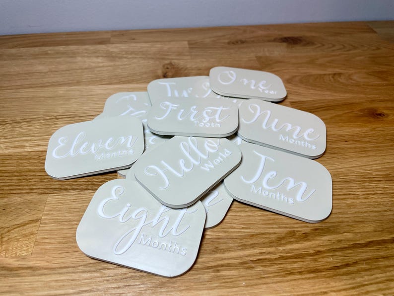 May include: A set of 11 beige square cards with white text. The cards feature different phrases and numbers, such as "One Month", "First", "Hello World", "Eight Months", "Eleven Months", "Nine Months", "Ten Months", and "Two Months".