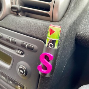 Pu&ograve; includere: Un interno di auto nero con una lettera "S" rosa attaccata a un supporto nero. Un tubo di balsamo per labbra verde e bianco &egrave; nel supporto.