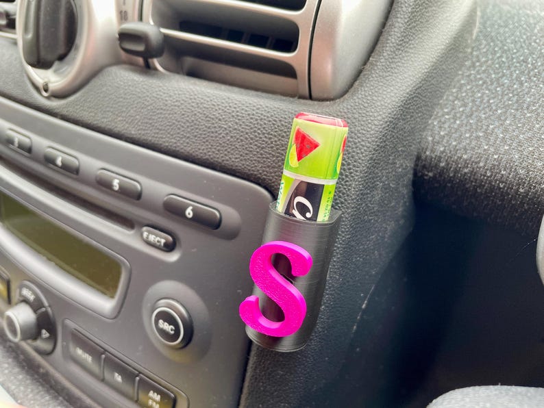 Pu&ograve; includere: Un interno di auto nero con una lettera "S" rosa attaccata a un supporto nero. Un tubo di balsamo per labbra verde e bianco &egrave; nel supporto.