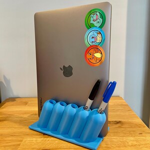 May include: A silver laptop with three circular character stickers vertically aligned on the back, held upright by a light blue plastic desk organizer featuring five cylindrical slots, containing one black and one blue marker pen.