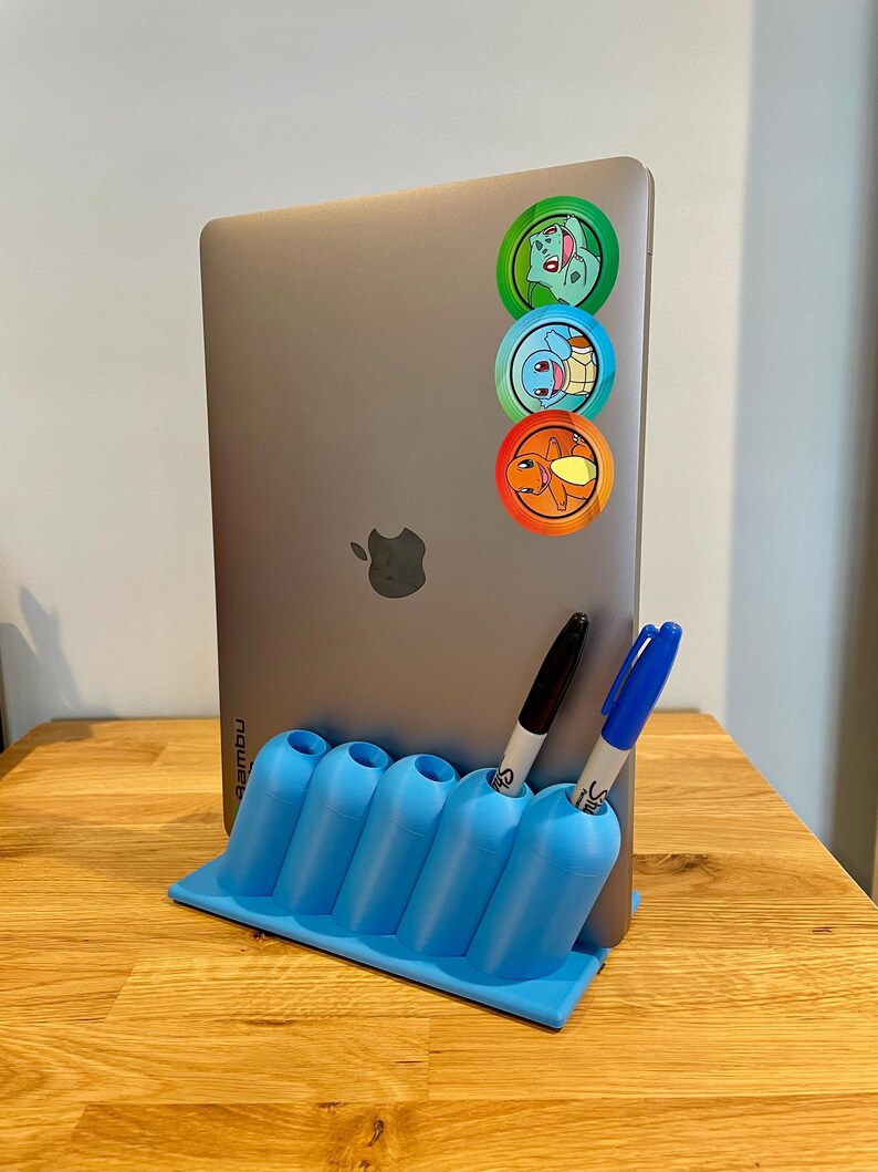 May include: A silver laptop with three circular character stickers vertically aligned on the back, held upright by a light blue plastic desk organizer featuring five cylindrical slots, containing one black and one blue marker pen.