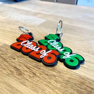 May include: Two keychains with the text "Class of 2026". One keychain is red and black, the other is green and black. The text "Class of" is white. The keychains are on a wooden surface.