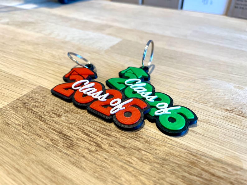 May include: Two keychains with the text "Class of 2026". One keychain is red and black, the other is green and black. The text "Class of" is white. The keychains are on a wooden surface.