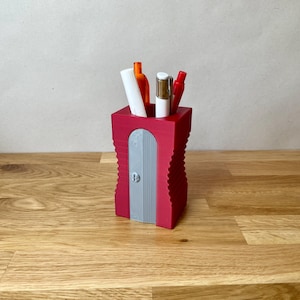 K&ouml;nnte beinhalten: Ein roter Plastik-Bleistifthalter mit integriertem Bleistiftspitzer. Der Halter hat ein geripptes Design und ist mit Stiften und Bleistiften gef&uuml;llt.