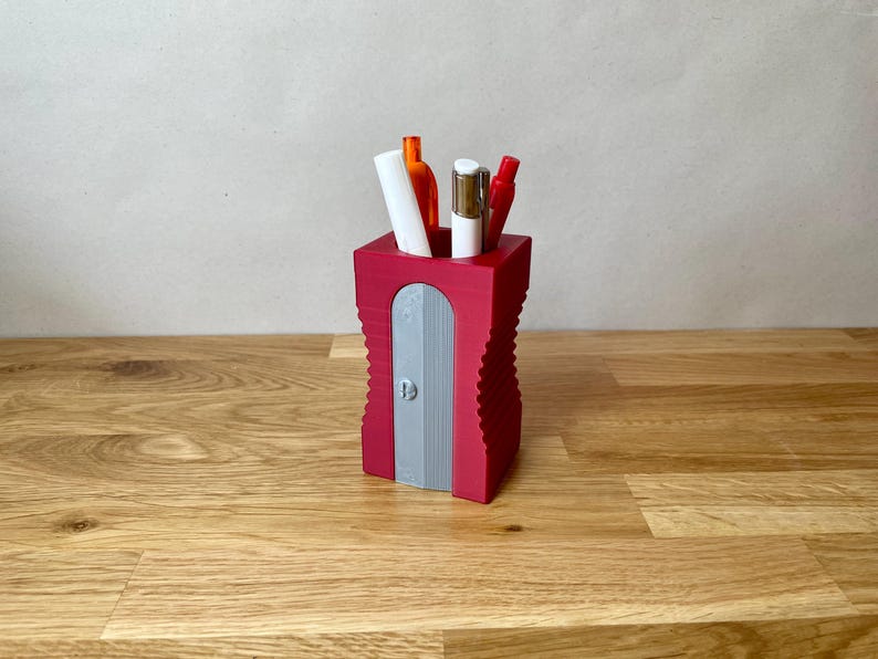 K&ouml;nnte beinhalten: Ein roter Plastik-Bleistifthalter mit integriertem Bleistiftspitzer. Der Halter hat ein geripptes Design und ist mit Stiften und Bleistiften gef&uuml;llt.