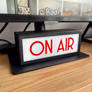 May include: A black rectangular "ON AIR" sign with red lettering, illuminated by a white light. The sign sits on a black base, placed on a wooden surface, and is positioned below a computer monitor.