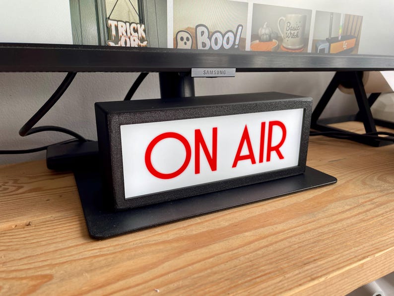 May include: A black rectangular "ON AIR" sign with red lettering, illuminated by a white light. The sign sits on a black base, placed on a wooden surface, and is positioned below a computer monitor.