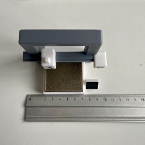 Pu&ograve; includere: Un oggetto grigio e bianco stampato in 3D con una base rettangolare e un telaio grigio. Un righello &egrave; posto accanto all'oggetto, mostrando le misure in centimetri. L'oggetto sembra essere uno strumento o una piccola macchina.