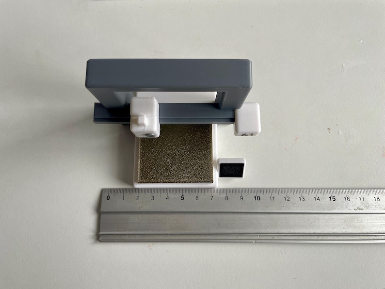 Pu&ograve; includere: Un oggetto grigio e bianco stampato in 3D con una base rettangolare e un telaio grigio. Un righello &egrave; posto accanto all'oggetto, mostrando le misure in centimetri. L'oggetto sembra essere uno strumento o una piccola macchina.