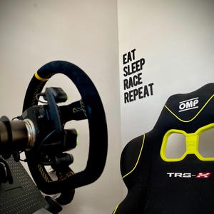 May include: A racing simulator setup featuring a black steering wheel with yellow accents and a black racing seat with yellow trim. The wall displays the text "EAT SLEEP RACE REPEAT".