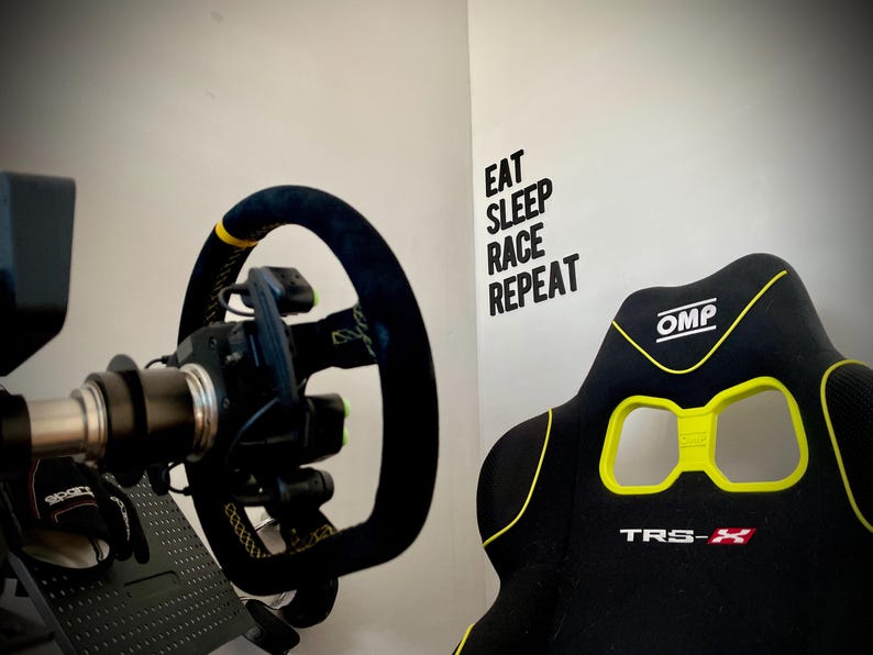 May include: A racing simulator setup featuring a black steering wheel with yellow accents and a black racing seat with yellow trim. The wall displays the text "EAT SLEEP RACE REPEAT".