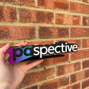 May include: A black and white "perspective" sign with a gradient of purple, blue, and black. The sign is held in front of a red brick wall.