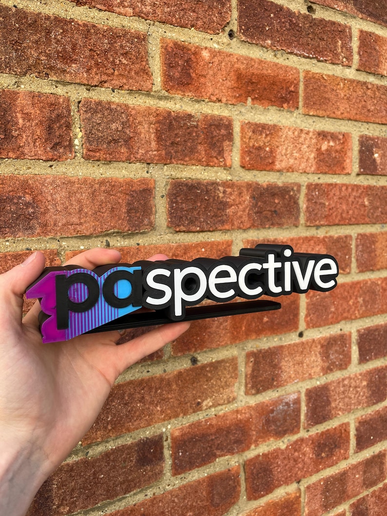 May include: A black and white "perspective" sign with a gradient of purple, blue, and black. The sign is held in front of a red brick wall.
