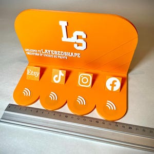 May include: An orange acrylic display stand with the text "WELCOME TO LAYEREDSHAPE CREATORS OF UNIQUE 3D PRINTS" and the initials "LS". It features icons for Etsy, TikTok, Instagram, and Facebook. A ruler is placed below the stand.