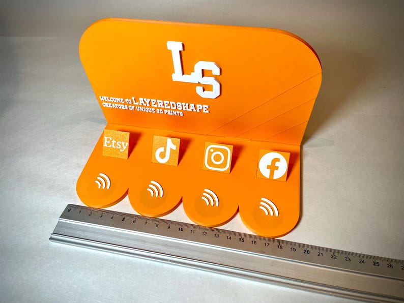 May include: An orange acrylic display stand with the text "WELCOME TO LAYEREDSHAPE CREATORS OF UNIQUE 3D PRINTS" and the initials "LS". It features icons for Etsy, TikTok, Instagram, and Facebook. A ruler is placed below the stand.