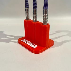 Op de afbeelding: Een rode 3D-geprinte darthouder met de naam "JOSHUA" in witte letters. Drie darts met zilveren en blauwe details zijn in de houder geplaatst. De houder staat op een wit oppervlak.