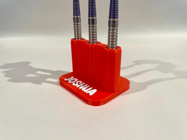 Op de afbeelding: Een rode 3D-geprinte darthouder met de naam "JOSHUA" in witte letters. Drie darts met zilveren en blauwe details zijn in de houder geplaatst. De houder staat op een wit oppervlak.