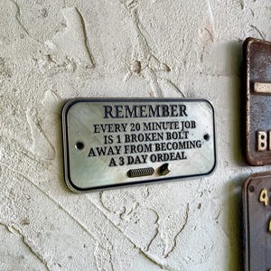 May include: A silver metal sign with black text that reads "Remember Every 20 minute job is 1 broken bolt away from becoming a 3 day ordeal."