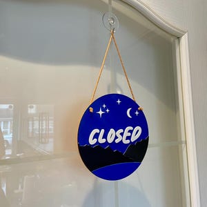 May include: A round blue and black sign with white stars and a crescent moon hanging on a glass door. The sign says "CLOSED" in white letters.