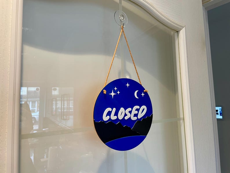 May include: A round blue and black sign with white stars and a crescent moon hanging on a glass door. The sign says "CLOSED" in white letters.
