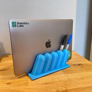 May include: A light blue 3D-printed plastic stand holding a silver laptop with a Bambu Lab logo and an Apple logo. The stand features five cylindrical slots, two of which contain a black marker and a blue marker, providing a stable vertical base.