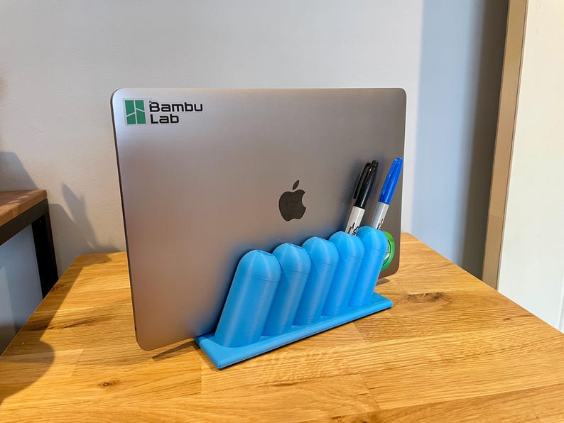 May include: A light blue 3D-printed plastic stand holding a silver laptop with a Bambu Lab logo and an Apple logo. The stand features five cylindrical slots, two of which contain a black marker and a blue marker, providing a stable vertical base.