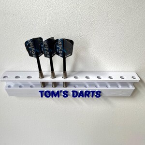 May include: A white dart holder with three darts. The darts have black shafts and blue and black flights. The holder has the text "TOM'S DARTS" in blue.