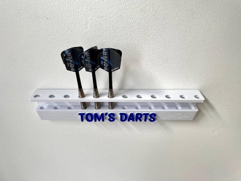 May include: A white dart holder with three darts. The darts have black shafts and blue and black flights. The holder has the text "TOM'S DARTS" in blue.