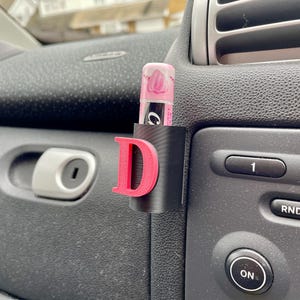 Pu&ograve; includere: Un accessorio per auto nero con una lettera "D" rosa e un porta-tubo per balsamo per labbra. Il tubo di balsamo per labbra &egrave; rosa e bianco con la parola "Carmex" stampata su di esso.