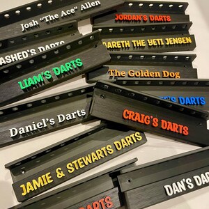 May include: A collection of black dart holders with various names in colorful 3D lettering. Names include "Josh 'The Ace' Allen", "Jordan's Darts", "Liam's Darts", "Daniel's Darts", "Craig's Darts", and others. The holders have a slotted design.
